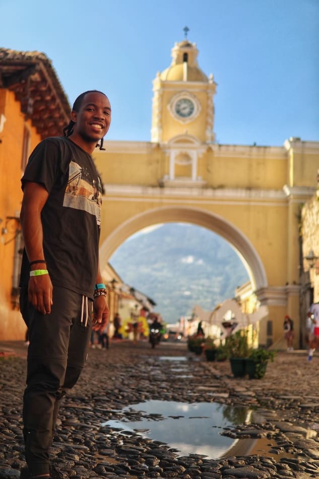 Black male budget traveler standing on cobble stone street in Guatemala with yellow building behind him