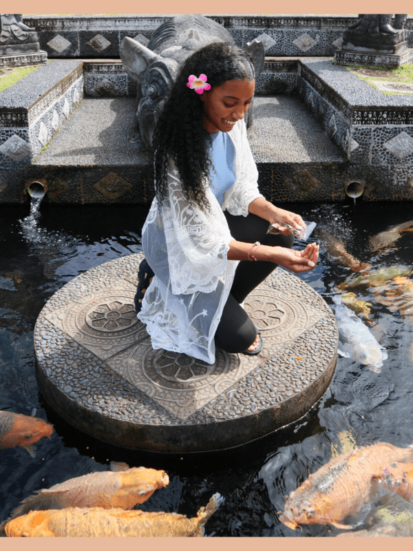 black female budget traveler squatting down feeding the large orange and silver fish in the water at tirta gangga water palace. She has long curly hair with a pink flower in her hair, a blue top, black leggings, black sandals, and a white cover up. She is smiling, looking down.