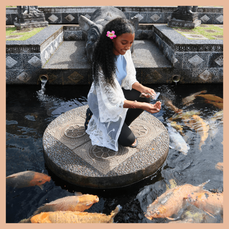 black female budget traveler squatting down feeding the large orange and silver fish in the water at tirta gangga water palace. She has long curly hair with a pink flower in her hair, a blue top, black leggings, black sandals, and a white cover up. She is smiling, looking down.