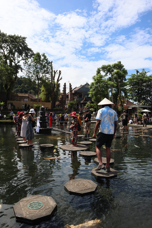 black male in blue and white shirt, black shorts, sneakers, and traditional hat walking on the path of water pillars at tirta gangga in bali with large coi fish swimming beneath him