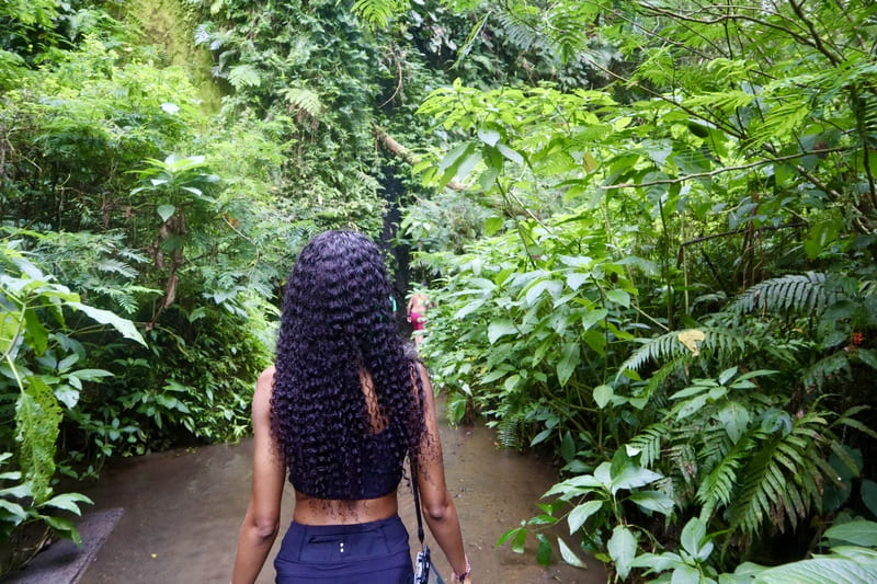 black traveler walking along the path to tukad cepung. she has long curly hair and is walking with green trees on both sides of her.