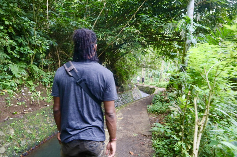 black traveler walking along the path to tukad cepung. there is a curved waterway to the left of him, as he walks along the sidewalk with green trees beside him