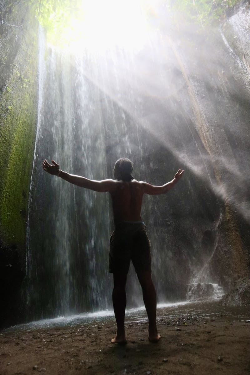 black traveler standing in front of tukad cepung waterfall with sun rays shining down on him in bali