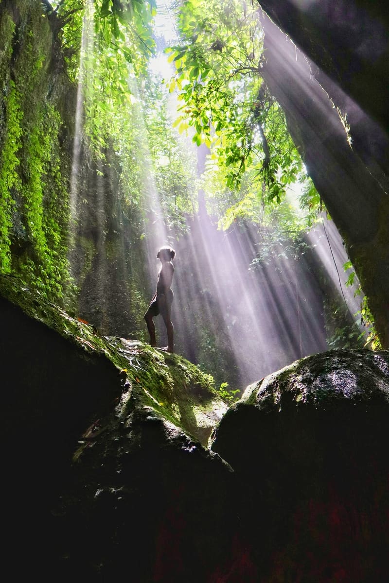 black traveler standing on large rock with sun rays shining down on him in tukad cepung in bali
