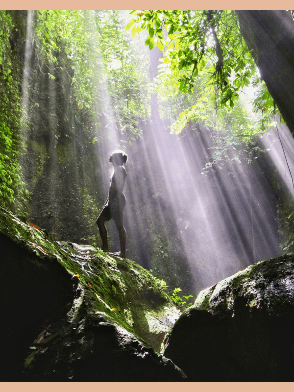 black traveler standing on top of large, green-mossed covered rock with sun rays shining down on him surrounded by greenery at tukad cepung in bali