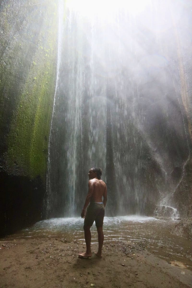 black traveler standing in front of tukad cepung waterfall with sun rays glistening over him and the waterfall. his back is to the camera, and his face is slightly turned to the camera.
