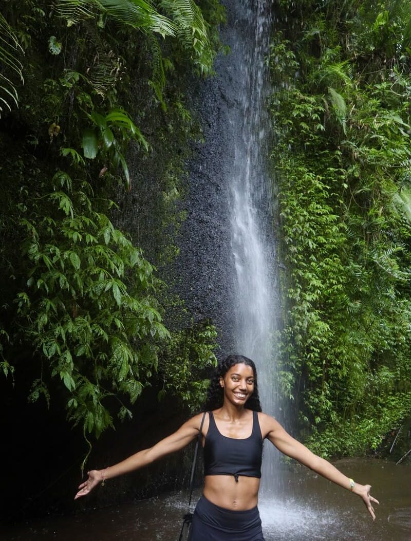 black traveler standing in front of the waterfall on the other side of tukad cepung in bali. she has her arms out to the sides and smiling at the camera.
