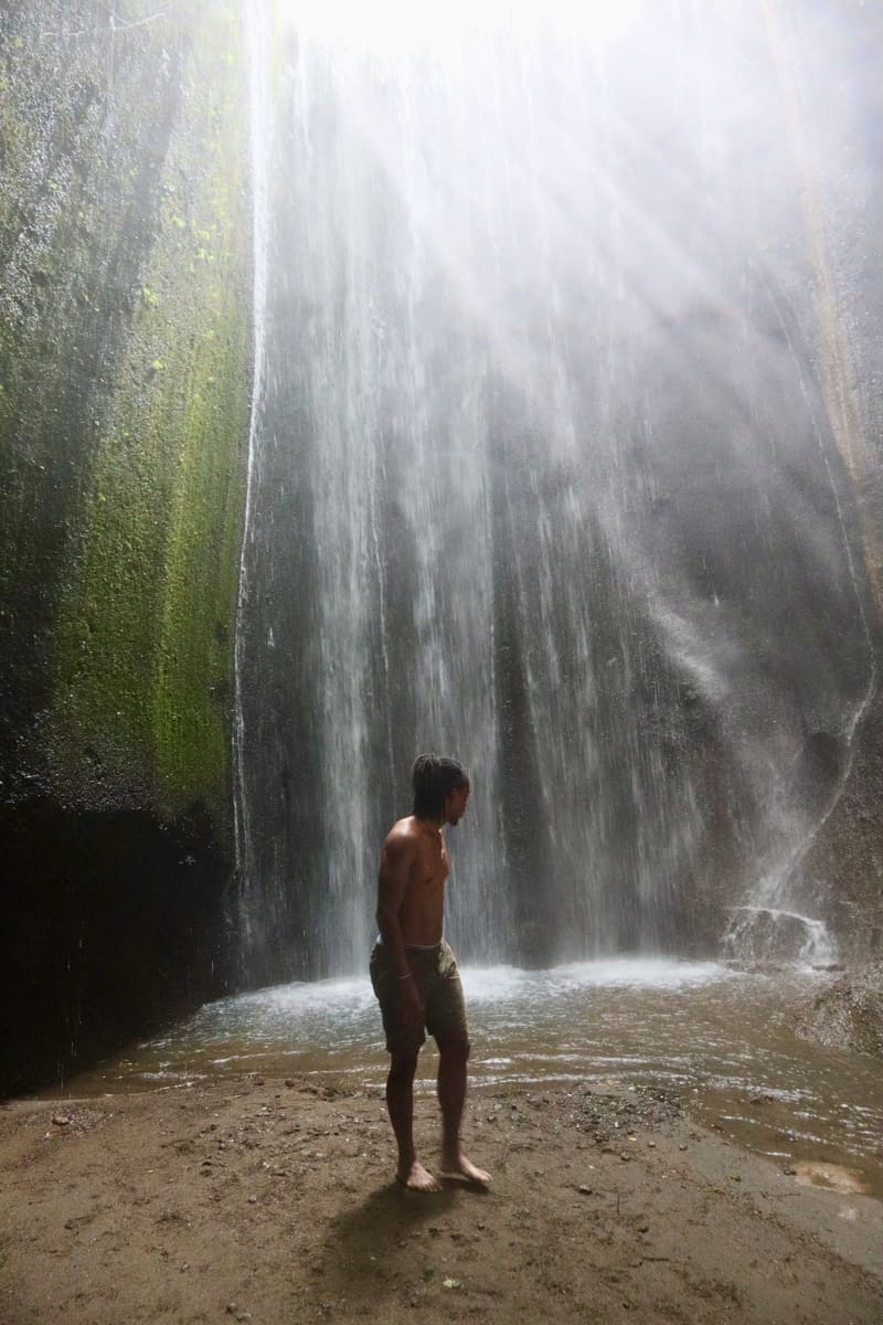 black traveler standing in front of tukad cepung waterfall looking back at the waterfall as he walks away from it. The sun rays are glistening over the waterfall.