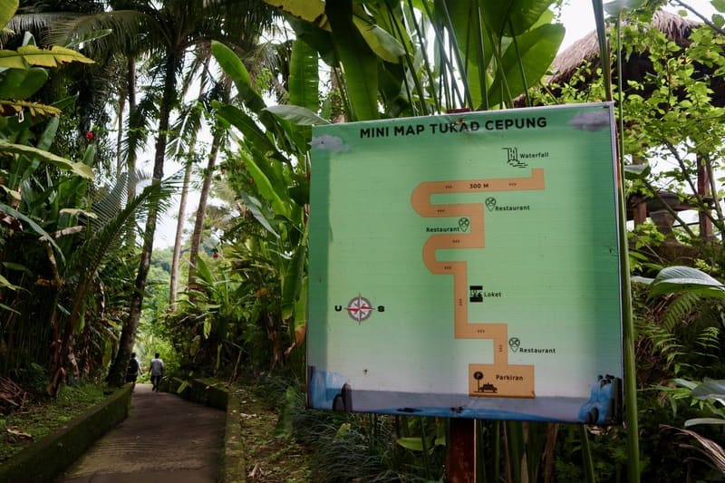 mini map of tukad cepung highlighting where the parking lot, restaurants, and waterfall are located. the map is hung up along the green trees in front of the path to tukad cepung