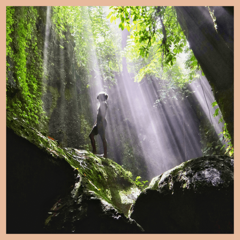 black traveler standing on top of large, green-mossed covered rock with sun rays shining down on him surrounded by greenery at tukad cepung in bali
